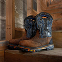 A pair of brown leather western work boots with blue and black embroidered shafts, resting on a wooden step in a rustic setting.