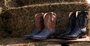 Four western cowboy boots displayed on a wooden surface in front of a large bale of hay. Two boots are brown with dark blue crocodile-embossed lower sections, and two are dark blue with light tan crocodile-embossed lower sections.