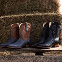 Two pairs of western cowboy boots displayed side by side on a wooden platform in front of stacked hay bales. The left pair features a brown leather upper with tan western embroidery and a dark blue textured lower shaft.