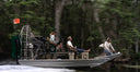 A small motorized airboat with four people traveling on water through a dense forest. The boat has a large rear fan, a green cabin area, and an orange flag flying at the back.