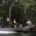 A gray motorized airboat with three people on board moves across calm water, with a dense green forest blurred in the background.