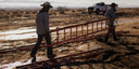 Two workers carry a long red metal ladder frame across a dry, muddy outdoor landscape near a white pickup truck in the background.