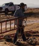 A man in a work hat and plaid shirt stands in a dusty outdoor setting near a red metal fence, holding ropes and chains. A white pickup truck is visible in the background.