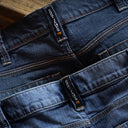 Close-up of dark blue denim jeans showing the waistband, belt loops, front pockets, and a branded fabric tab with white text and a small orange logo.