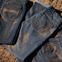 Close-up of folded blue denim jeans laid on a dirt surface, showing the back pockets and contrast stitching.