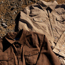 Two western-style button-up shirts laid flat on a gravel surface. The shirt in the foreground is brown and black checkered, while the shirt behind it is a light tan with a subtle geometric pattern.