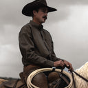 A man wearing a dark brown cowboy hat and a brown patterned long-sleeve shirt sits on a horse, holding a coiled rope under a cloudy gray sky.