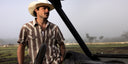 A man wearing a white cowboy hat and a brown and beige striped short-sleeve button-up shirt stands beside a vehicle in an outdoor field with a hazy background.