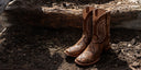 A pair of brown western cowboy boots with a snakeskin pattern and decorative stitching, standing on dark soil next to a large log outdoors.