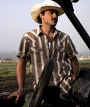 A man wearing a white cowboy hat and a brown and beige striped short-sleeve western shirt stands in a field next to a vehicle.