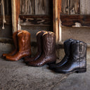 Three exotic skin cowboy boots in brown, tan, and black are lined up on a rustic stone floor in front of weathered wooden doors.